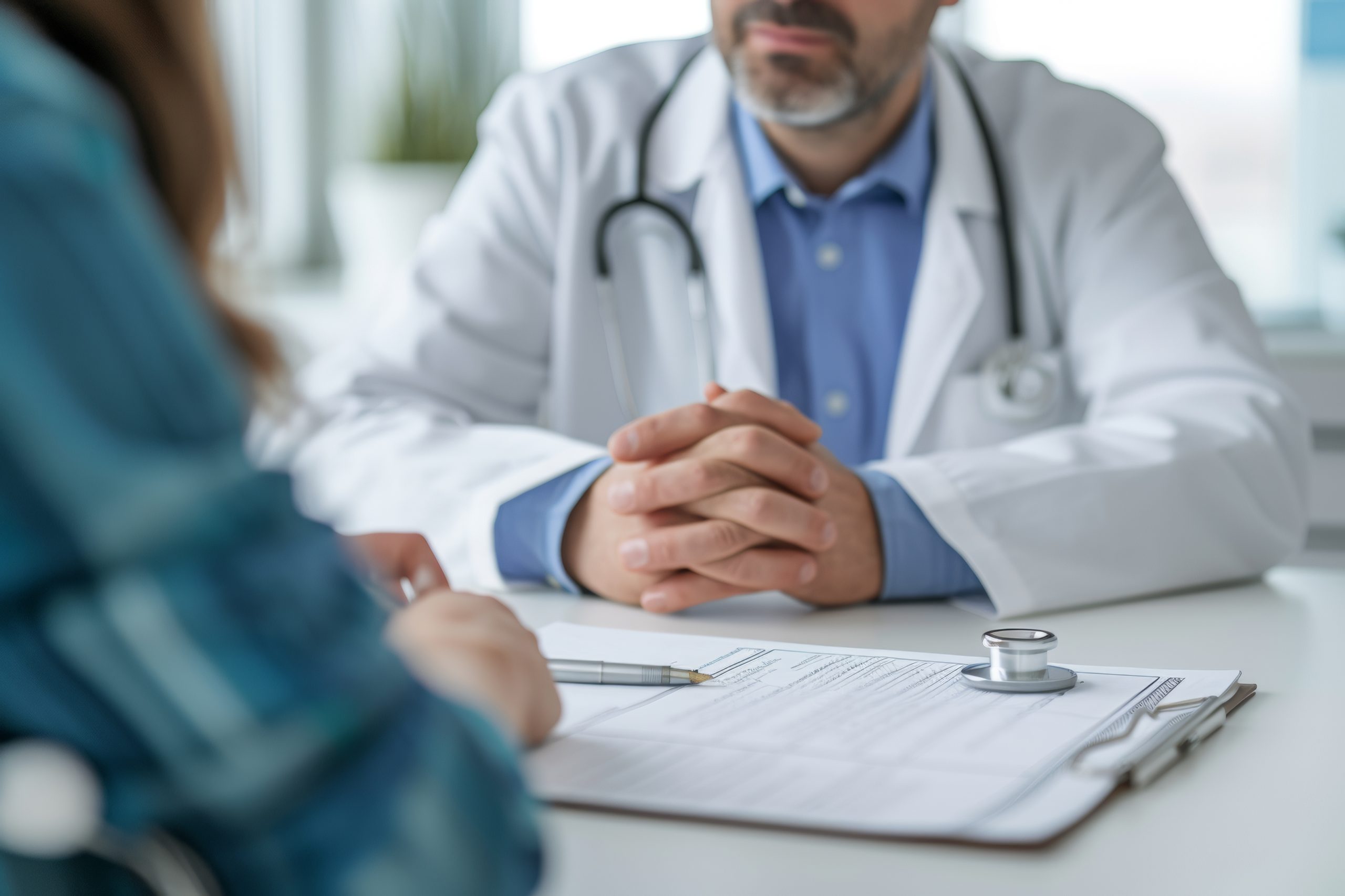 Doctor in a white coat consulting with a patient, focus on clasped hands and medical paperwork on the desk
